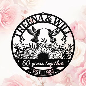May include: Black metal wall art featuring two hummingbirds facing each other above a floral design. The circular piece includes the names "TREENA & WILL", "60 years together", and "EST. 1965". The background is a soft pink with rose blossoms.