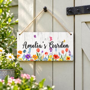 Puede incluir: Un letrero rectangular de madera con fondo de tablones blancos, que presenta el texto negro Amelia's Garden. El diseño incluye coloridas ilustraciones en acuarela de varias flores y pequeñas mariposas en el borde inferior.