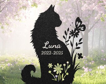 May include: Black metal cat silhouette garden stake with the name "Luna" and dates 2022-2025. The cat is surrounded by floral and butterfly cutouts. The background features a blurred garden scene with pink flowers.