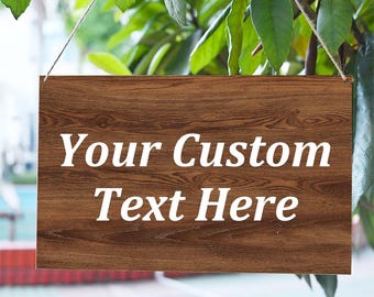 May include: A rectangular wooden sign with the text "Your Custom Text Here" in white. The sign is suspended by a thin string and has a wood grain texture. Green leaves are visible in the background.