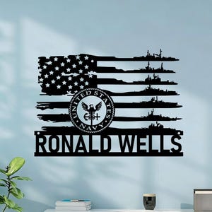 May include: Black metal wall art featuring a stylized American flag design with the United States Navy emblem. The flag includes the name "RONALD WELLS" at the bottom and silhouettes of naval ships.