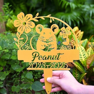 Könnte beinhalten: Ein gelber Gartenstecker mit einem ausgeschnittenen Design, das eine Maus, Blumen und das Wort "Peanut" mit den Jahren 2023-2025 zeigt. Der Stecker wird vor einem grünen, belaubten Hintergrund gehalten.