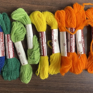 May include: A collection of colorful embroidery floss skeins in shades of blue, green, yellow, orange, and red. Each skein is labeled with "Needlepoint & Crewel Wool by Bucilla". The skeins are arranged in a row on a wooden surface.