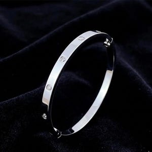 May include: A silver-colored bangle bracelet with four small, round, clear gemstones evenly spaced along the top. The bracelet has a smooth, polished finish and a hinged clasp. The background is a dark, textured fabric.