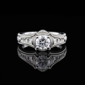 May include: A silver engagement ring with a large, round, clear gemstone at the center. The ring features intricate detailing and smaller, rectangular gemstones on the sides. The ring is set against a black background, with its reflection visible.