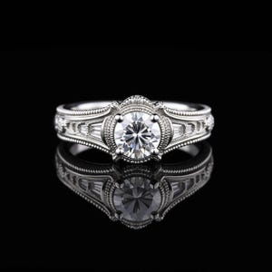 May include: A silver engagement ring with a large, round, clear gemstone at the center. The ring features intricate detailing and smaller, rectangular gemstones on the sides. The ring is set against a black background, with its reflection visible.