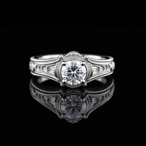 May include: A silver engagement ring with a large, round, clear gemstone at the center. The ring features intricate detailing and smaller, rectangular gemstones on the sides. The ring is set against a black background, with its reflection visible.
