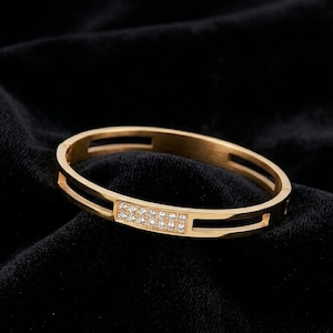May include: A gold-colored bangle bracelet with a rectangular section of small, clear gemstones. The bracelet has a simple, elegant design with cut-out sections on the sides. The bracelet is displayed on a black surface.
