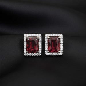 May include: A pair of stud earrings featuring rectangular, deep red gemstones surrounded by a halo of small, clear stones. The earrings are set against a black background, highlighting their sparkle and design.