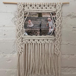 May include: Cream-colored macrame photo holder with a wooden dowel at the top. The woven design frames a photograph of two children on a park bench. The macrame has a fringe at the bottom, adding a decorative touch to this wall hanging.