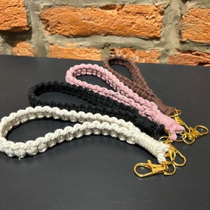 Puede incluir: Cuatro correas de muñeca de macramé en crema, negro, rosa y marrón. Cada correa está en bucle y unida a un cierre dorado. Las correas están dispuestas sobre una superficie oscura con una pared de ladrillo al fondo.