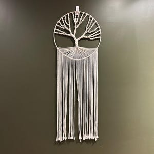 May include: A white macrame wall hanging featuring a tree design. The tree is within a circular frame, with long, vertical fringe hanging below. The wall hanging is suspended against a dark green wall.