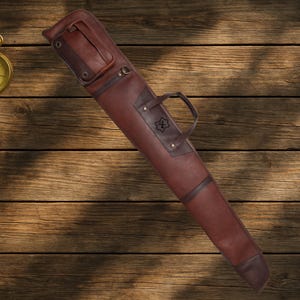 Puede incluir: Un estuche de rifle de cuero marrón con asa y costuras decorativas. El estuche tiene forma rectangular con un extremo redondeado y cierre de solapa. Está hecho de cuero marrón oscuro y tiene un asa para transportarlo.