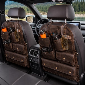 May include: Two dark brown leather car seat organisers attached to the back of front vehicle seats. Each organiser features multiple pockets, including vertical sleeves for bottles, smaller pouches for pens or tools, and wide horizontal compartments.