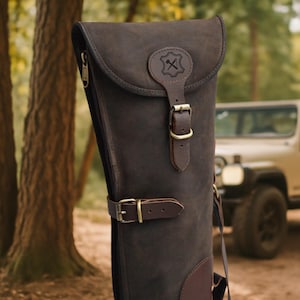 Puede incluir: Un estuche para rifle de cuero marrón oscuro con cierre de solapa y hebilla. El estuche tiene una cremallera en el lateral y un pequeño emblema en la solapa. El fondo muestra un bosque y un vehículo.