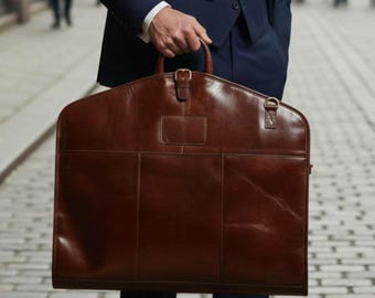 Könnte beinhalten: Eine braune Leder-Kleidersack mit einem oberen Griff und einem Schnallenverschluss. Die Tasche hat eine rechteckige Form mit einer Vordertasche. Eine Person in einem marineblauen Anzug hält sie.