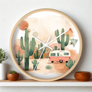 May include: A round wall clock with a light wood frame featuring a watercolor illustration of a desert landscape with green cacti, a pink and white camper trailer, and abstract geometric shapes in shades of orange, tan, and cream on a white background.