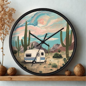 May include: A round wall clock with a black frame featuring a desert landscape illustration. The scene depicts a vintage white camper with blue stripes, tall green saguaro cacti, and distant mountains under a swirling pink and blue sky.