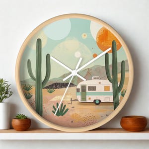 May include: A round wall clock with a light wood frame featuring a desert landscape illustration. The scene depicts a vintage white camper trailer, tall green saguaro cacti, and distant mountains under a pale blue sky with orange sun circles.