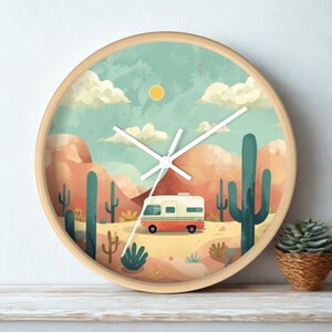 Könnte beinhalten: Eine runde Wanduhr mit hellem Holzrahmen und einer Wüstenlandschaft-Illustration. Das Motiv zeigt einen Wohnwagen, hohe grüne Kakteen, orangefarbene Felsformationen und eine gelbe Sonne unter einem hellblauen Himmel mit weißen Wolken.