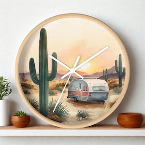 Könnte beinhalten: Eine runde Wanduhr mit hellem Holzrahmen und einer Wüstenlandschaft-Illustration. Die Szene zeigt einen Vintage-Wohnwagen in einer sandigen Gegend mit hohen grünen Saguaro-Kakteen, kleineren Wüstenpflanzen und fernen Bergen unter einem sanften Sonnenuntergangshimmel.
