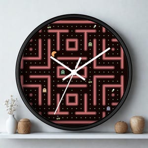 May include: A round black wall clock featuring a Pac-Man maze design in shades of pink and black. The clock has white hands and a black frame. The clock is a decorative item.
