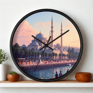May include: A round wall clock with a black frame featuring a printed image of a mosque with multiple domes and tall minarets at sunset. The scene includes palm trees, a waterfront promenade with people, and a calm body of water in the foreground.