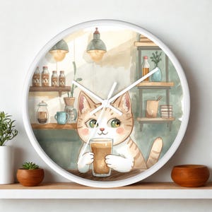 Op de afbeelding: Een ronde wandklok met een witte lijst voorzien van een aquarelillustratie van een lichtoranje cyperse kat die een glas koffie vasthoudt. De achtergrond toont een café-interieur met planken, flessen, hanglampen en een groene muur.