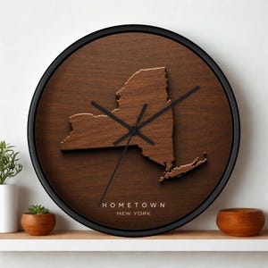 Può includere: Un orologio da parete rotondo con cornice nera e quadrante in legno marrone scuro. Un ritaglio in legno tridimensionale in rilievo dello stato di New York è centrato sul quadrante. Il testo HOMETOWN NEW YORK è stampato in bianco sotto la mappa.