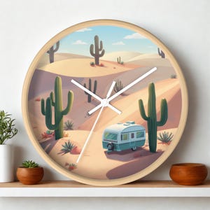 Könnte beinhalten: Eine runde Wanduhr mit hellem Holzrahmen und einer Wüstenlandschaft-Illustration. Die Szene zeigt einen hellblauen und weißen Wohnwagen, der zwischen grünen Saguaro-Kakteen auf Sanddünen unter einem blassblauen Himmel mit weißen Wolken steht.