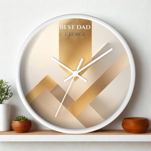 May include: A round wall clock with a white frame and a geometric face featuring shades of beige, cream, and metallic gold. The text BEST DAD and GEORGE is printed in white and dark grey at the top. It has three white clock hands at the centre.
