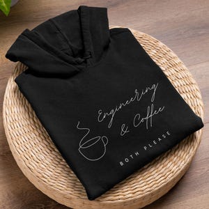 Pode incluir: Moletom preto com a frase "Engineering & Coffee BOTH PLEASE" em escrita branca. Um desenho de linha branca de uma xícara de café. O moletom está dobrado sobre um tapete redondo tecido.