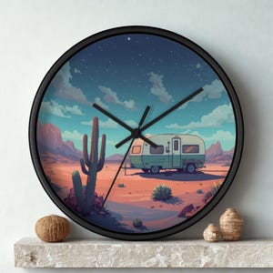 May include: Black-framed wall clock featuring a desert landscape with a green and white camper, cacti, and mountains under a starry sky. The clock hands are black, and the clock is displayed on a white surface.