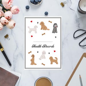 May include: A white, rectangular "Health Records" notebook featuring illustrations of various dog breeds, paw prints, and bone graphics. The notebook is surrounded by office supplies, flowers, and a cup of coffee on a marble surface.