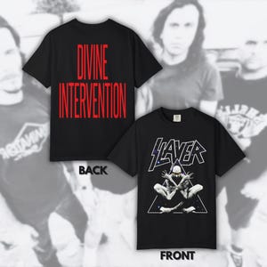 Könnte beinhalten: Ein schwarzes Kurzarm-T-Shirt mit einer Grafik des Bandnamens Slayer auf der Vorderseite, die eine sitzende Kreatur in einem weißen Dreieck zeigt. Auf der Rückseite steht der rote Text Divine Intervention in einer vertikalen Schriftart.