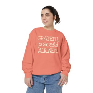 May include: A coral-colored sweatshirt with the words "Grateful, Peaceful, Aligned" printed in a cream-colored font. The long-sleeved top is paired with blue jeans. The sweatshirt is a casual, comfortable garment.
