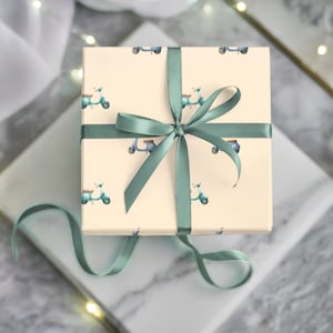 May include: A square gift box wrapped in cream-colored paper, patterned with small blue scooters. The box is tied with a sage green satin ribbon, forming a bow on top. The gift sits on a marble surface.