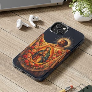 May include: A navy blue iPhone case featuring a religious image of a saint holding a banner with a depiction of the Virgin Mary surrounded by roses. The case is on a wooden surface with a laptop, earbuds, and a plant in the background.