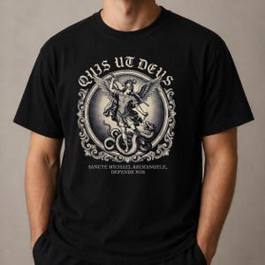 May include: Black t-shirt with a detailed monochrome illustration of a winged figure with a sword, fighting a serpent, within an ornate circular frame. The text "QUIS UT DEUS" and "SANCTE MICHAEL ARCHANGELE, DEFENDE NOS" are displayed above and below.