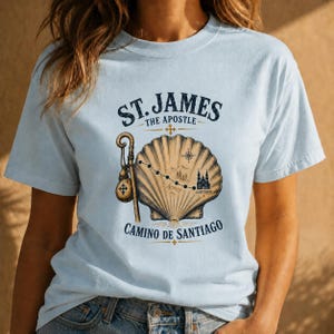 May include: A light blue t-shirt featuring a graphic of a scallop shell, a pilgrim staff with a gourd, and a map route leading to a cathedral. The text reads ST. JAMES THE APOSTLE and CAMINO DE SANTIAGO in dark blue lettering on the front.