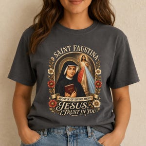May include: A charcoal grey t-shirt featuring a graphic print of Saint Faustina holding a diary, with an image of Jesus in the background. The text reads Saint Faustina, Apostle of Divine Mercy, and Jesus, I Trust In You, framed by floral accents.