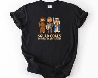 Op de afbeelding: Zwart T-shirt met een cartoon illustratie van St. Franciscus, St. Joan en St. Teresa. De tekst "SQUAD GOALS" staat boven de figuren, met hun namen eronder. Het T-shirt is gemaakt van een zacht materiaal.