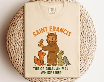May include: Beige t-shirt featuring a cartoon illustration of Saint Francis with a halo, holding a bird, surrounded by a fox, rabbit, and dove. The text "SAINT FRANCIS" and "THE ORIGINAL ANIMAL WHISPERER" are printed in orange and green.