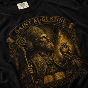 May include: Black t-shirt featuring a detailed gold illustration of Saint Augustine holding a staff and a flaming heart. The design includes the text "SAINT AUGUSTINE" and Latin phrases. The shirt has a Comfort Colors tag.