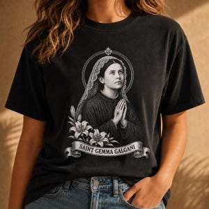 Peut inclure: Un t-shirt noir à manches courtes avec une impression graphique blanche représentant Sainte Gemma Galgani portant un voile, les mains jointes en prière, entourée de lys. Une bannière porte le texte SAINT GEMMA GALGANI.