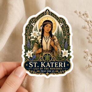 May include: A die-cut sticker featuring an illustration of St. Kateri Tekakwitha holding a wooden cross and white lilies. The design includes a turtle emblem, pine trees, and the text ST. KATERI LILY OF THE MOHAWKS PRAY FOR US in gold.