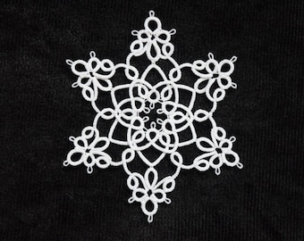 Op de afbeelding: Een witte, zespuntige sneeuwvlok-doily met ingewikkelde kanten details. Het delicate ontwerp heeft luspatronen en bloemachtige vormen, tegen een donkere, getextureerde achtergrond. Dit handgemaakte item is perfect voor winterdecoratie.