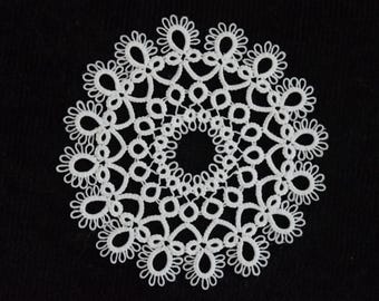 Op de afbeelding: Een witte, cirkelvormige doily met een ingewikkeld kanten ontwerp. De doily heeft een herhalend patroon van bloemen en druppelvormen, waardoor een delicate en symmetrische uitstraling ontstaat tegen een zwarte achtergrond. Handgemaakt item.