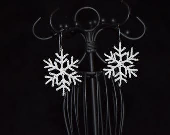 Op de afbeelding: Een paar witte sneeuwvlok oorbellen hangen aan een zwarte displaystandaard. De oorbellen zijn gedetailleerd met ingewikkelde patronen en de haken zijn zilverkleurig. De achtergrond is effen zwart, wat het delicate ontwerp van de sieraden benadrukt.