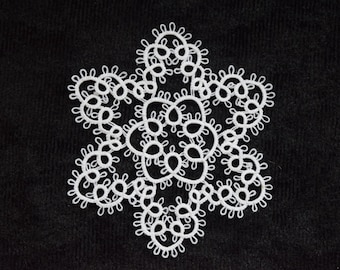 Op de afbeelding: Een witte, sneeuwvlokvormige doily tegen een zwarte achtergrond. De doily is gemaakt van delicate, ingeweven kant, met een symmetrisch ontwerp met afgeronde bloemblaadjes en een centraal patroon. De zwarte achtergrond zorgt voor een scherp contrast.